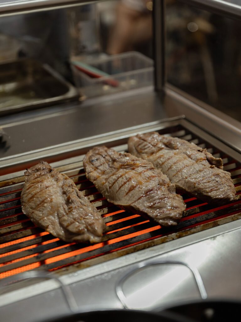 Several beef steaks grilling on a flame grill inside a stainless steel cooker, with an orange glow beneath the bars.