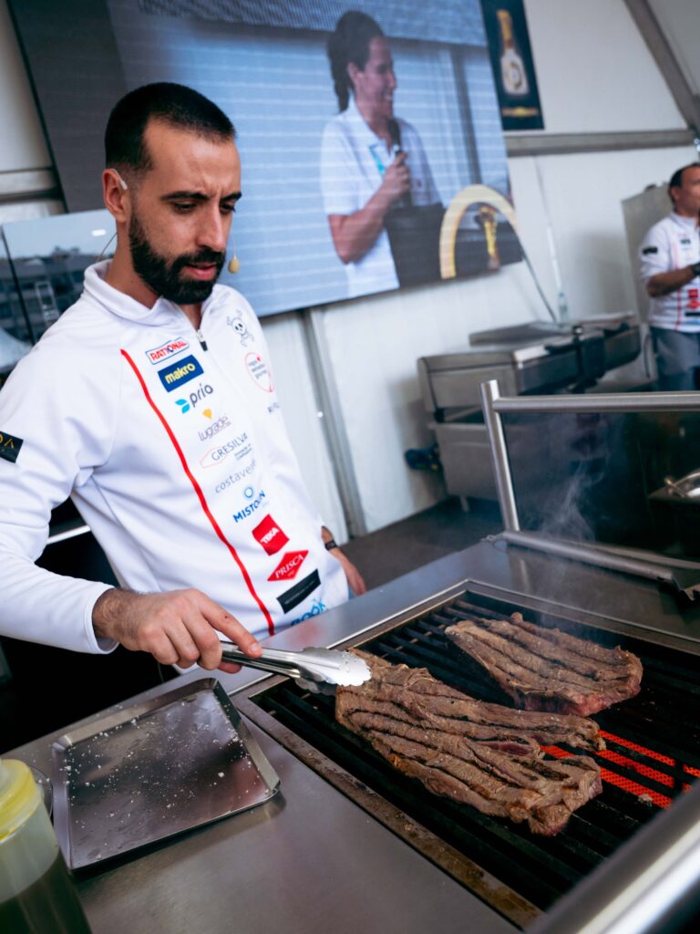 Chef wearing a white uniform and headset cooks sliced beef on a grilling station, turning meat with tongs as a large grill vents smoke in a professional kitchen.