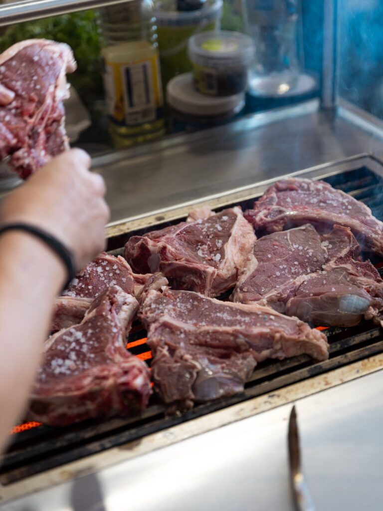 Person’s hand holding a salted ribeye over a hot grill, raw beef pieces on the grates with orange glow from embers in background