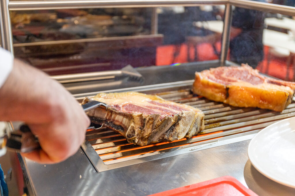 Meat chops grilling on a barbecue grill with flames underneath and a person tending the grill on the left.